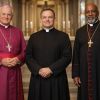 Three clergy members wearing cassocks standing together inside a church. The first clergy member wears a purple cassock, while the other two wear black cassocks with red trim, each adorned with crosses.