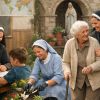 A group of nuns wearing traditional habits, interacting with children and an elderly woman. One nun is teaching children in a classroom setting, while another is helping in the garden. The nuns' caring roles exemplify the spiritual and service-oriented life they lead, in line with the traditions of the religious order.