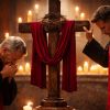 Two Catholic priests in red vestments, one kneeling in prayer and the other standing with his hand on a cross draped in red cloth, illuminated by candlelight during Good Friday services
